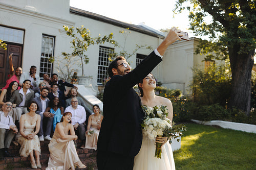 Bride and groom take a joyful wedding selfie with their enthusiastic guests