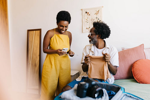 Happy mature couple smiling and chatting while packing clothes for romantic vacation in a bedroom