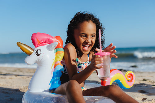 Happy girl enjoying a summer day at the beach with unicorn float and drink