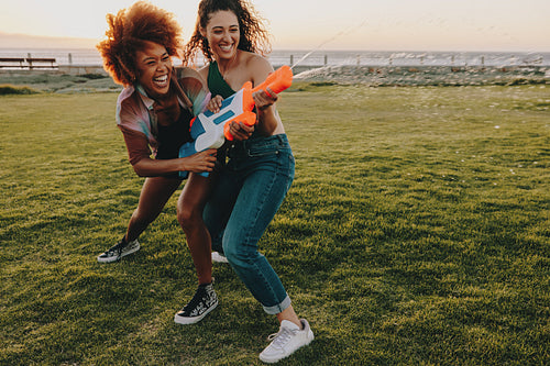 Two friends laugh and play with water guns in a city park at sunset, embodying fun and friendship in an outdoor atmosphere