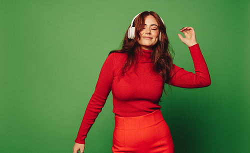 Cheerful young woman dancing to music with wireless headphones against vibrant green background