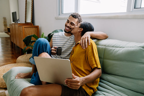 Gay couple relaxing together on a couch sharing a laptop and a coffee