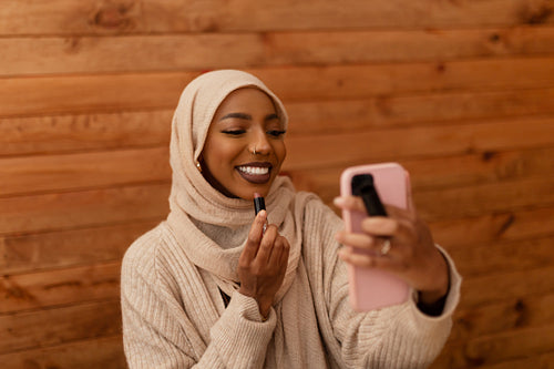 Stylish muslim woman using her smartphone as a mirror to apply l