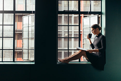 Woman sitting on window sill and reading book with coffee