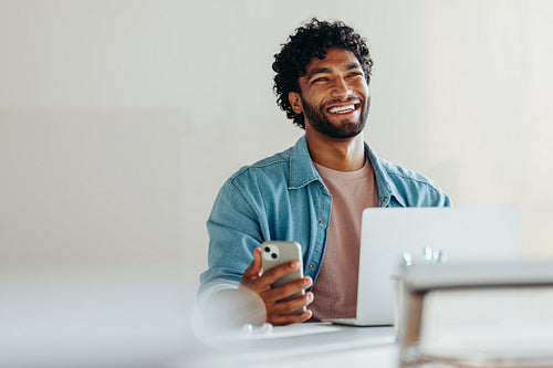 Happy young business man sitting with smartphone and laptop at desk
