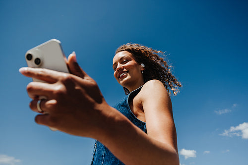Woman checks smartphone while listening to music outdoors under blue sky