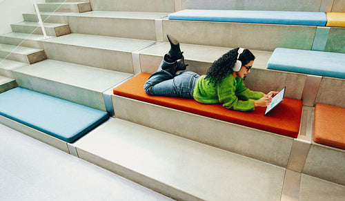 In a quiet library corner, the woman uses a tablet with headphones on indoor steps