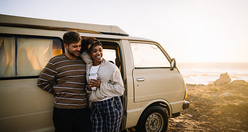 Couple smiles beside a camper van on a sunny beach during golden hour