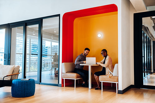 Two adults conversing and working at a shared table with a laptop
