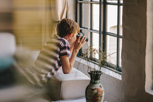Curious boy looking out the window with binocular