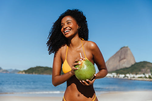 Joyful black woman in bikini enjoying a fresh coconut on a sunny day at the beach