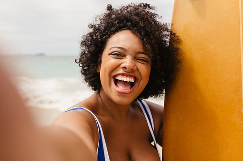 Happy woman smiling for a selfie at the beach, wearing a swimsuit and holding a surfboard