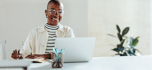 Portrait of a happy businesswoman working on a laptop in bright office with a notepad
