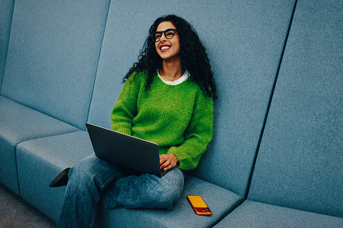 Smiling woman using laptop in public lounge area with smartphone