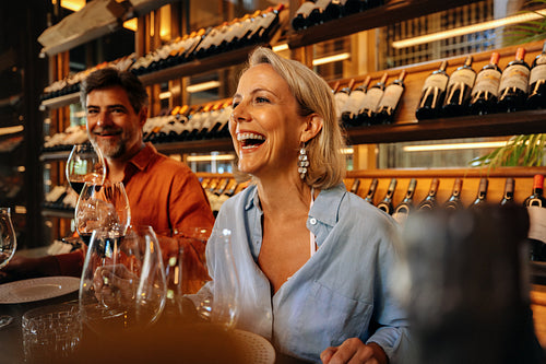 Mature woman laughing and enjoying wine with a man in front of a wine rack at a cozy restaurant