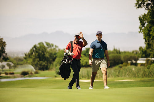 Sportsmen enjoying a sunny day on the golf course, walking together while carrying a golf bag and happily waving to friends