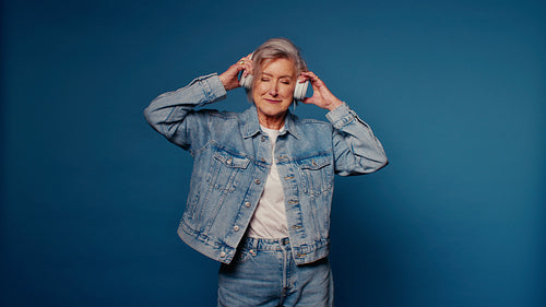 Happy senior woman enjoying music with headphones in a studio with blue backdrop