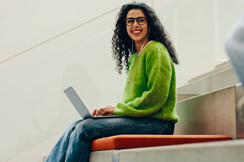 The smiling student studies on a laptop with headphones in library stair seating
