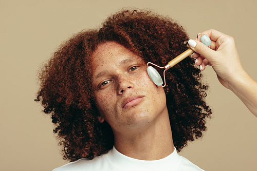 Portrait of a young man getting a jade roller massage on his freckled face