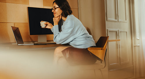 Woman works at desk on laptop while holding a coffee cup in home