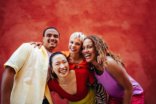 Young adult friends stand with arms around each other against a red wall outdoors