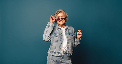 Playful senior woman peeking and smiling, casual denim outfit on blue background
