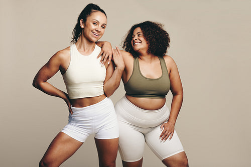 Athletic and proud, two female friends stand in gym wear showing their fit bodies