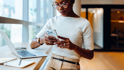 Businesswoman reading her smartphone while working on her laptop at a desk