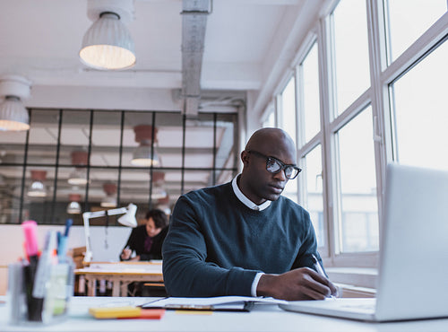African man writing notes while working on a laptop