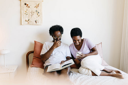 Happy mature couple laughing and relaxing together in bed while reading and using technology