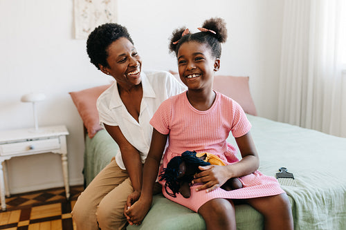 Happy young girl sitting with her mom and her doll on a bed
