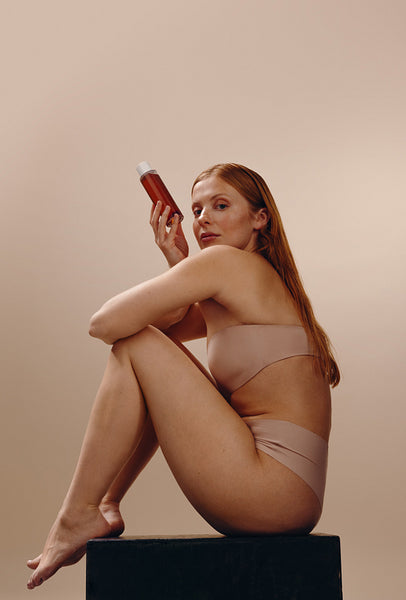 Woman holding skincare bottle while seated in neutral studio