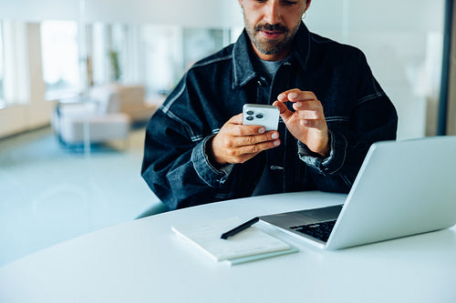 Man uses smartphone at desk with laptop in office