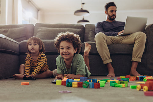 Man working on laptop sitting at home with kids playing on the floor