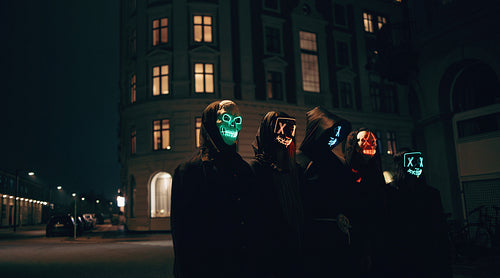 Friends in illuminated neon masks posing as mysterious figures on a dark city street for Halloween