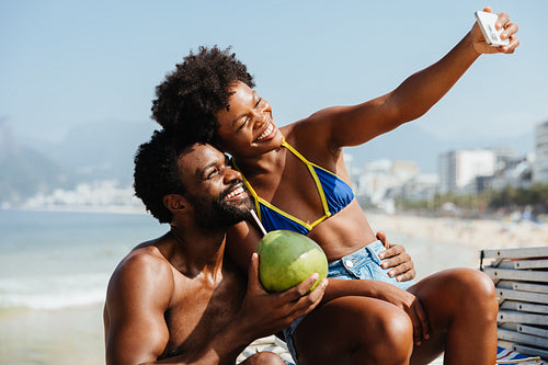 Joyful couple on honeymoon taking a beach selfie with coconut