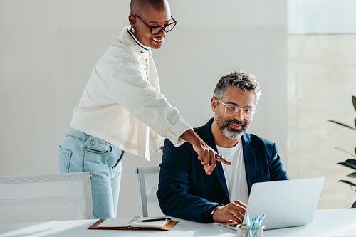 Happy young woman guiding colleague on laptop in bright office setting, teamwork and assistance concept