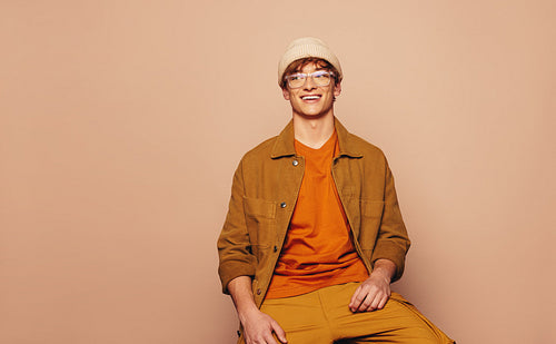 Cheerful young man with stylish urban style sitting and smiling in a vibrant peach background