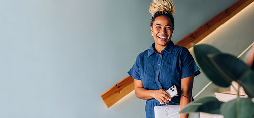 Smiling woman with blonde braided bun holding smartphone and tablet at staircase
