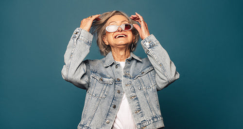 Playful senior woman in denim outfit and glasses smiling against blue studio background