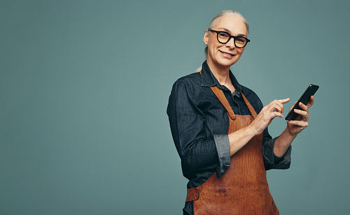 Cheerful mature goldsmith using a smartphone in a studio