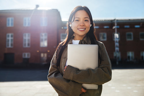 Smiling student holding laptop on sunny college campus