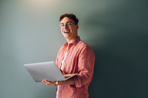 Young man smiling joyfully while holding a laptop against a plain background