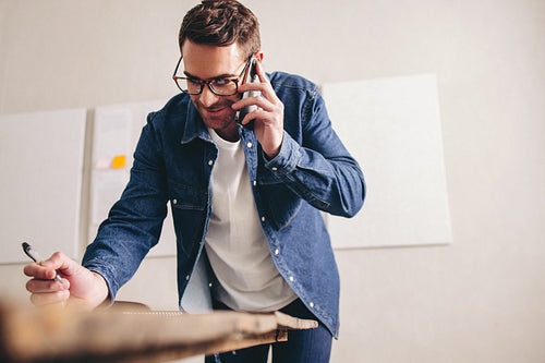 Cheerful businessman making notes during a phone call in his office
