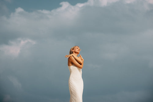 Joyful woman in white dress embracing herself under a moody cloudy sky