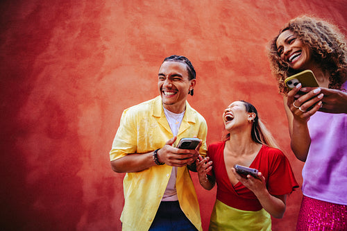 Young adult friends laugh over a smartphone together outdoors against a red wall