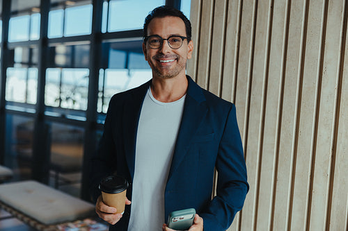 Man in business casual style stands with a phone and coffee in a modern office