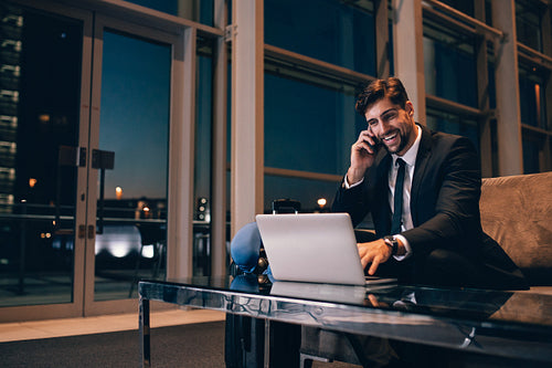 Smiling businessman with laptop talking on cellphone at the airport lounge