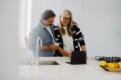 Woman watches as senior man uses a tablet in the kitchen