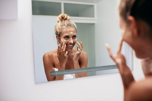 Happy young woman applying cream in bathroom